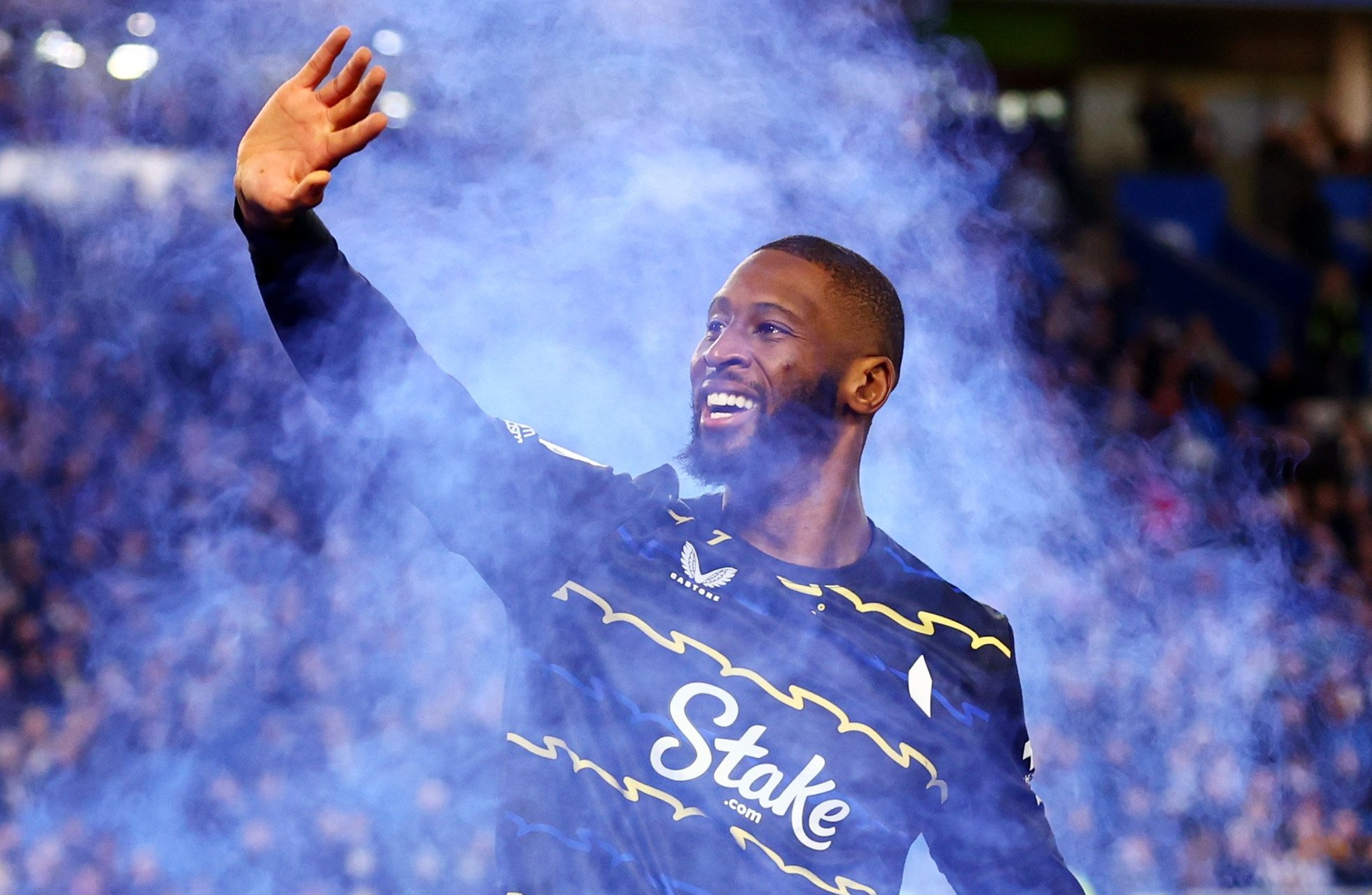Beto of Everton celebrates scoring a goal, raising his hand, surrounded by blue smoke on the football field.