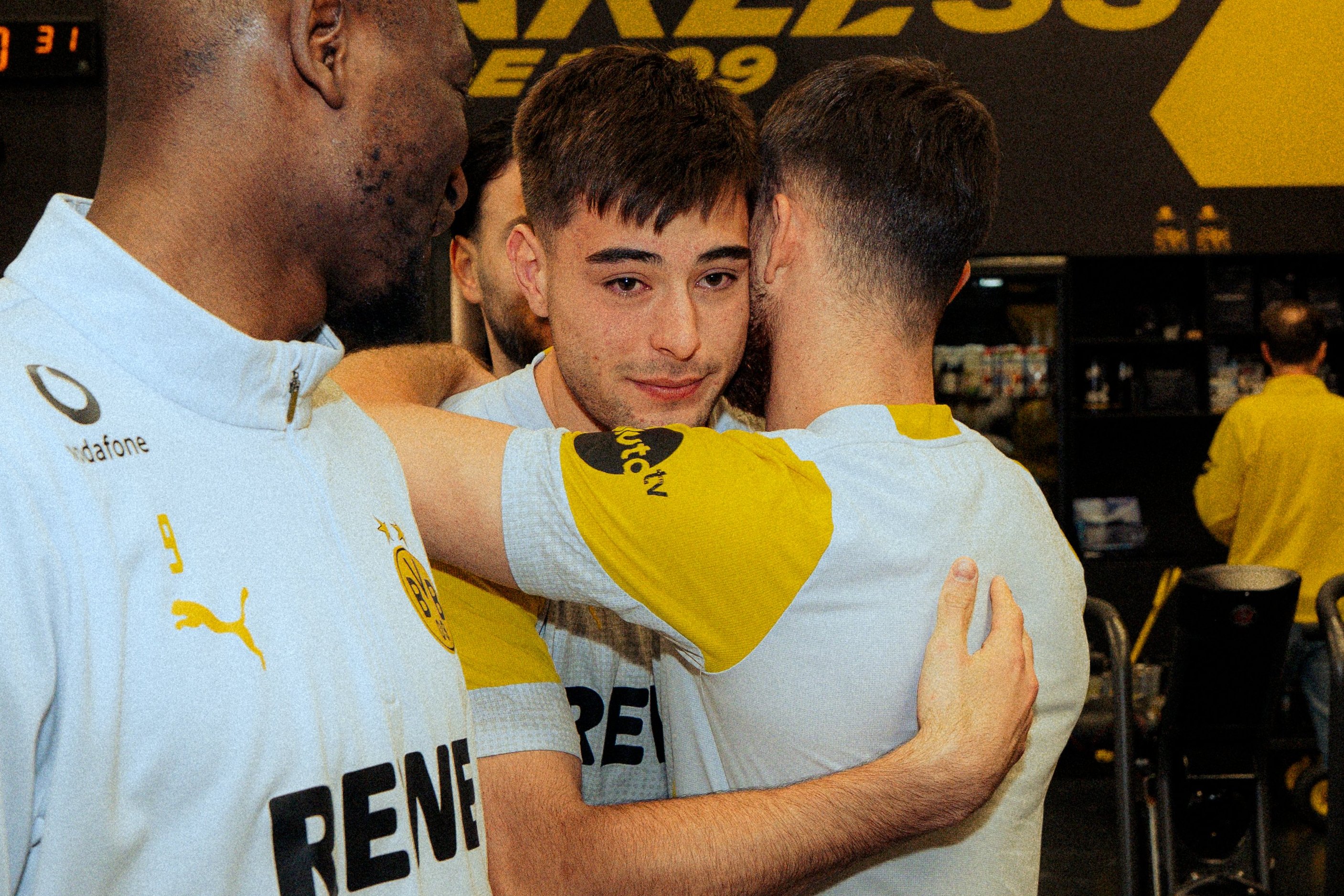Three men in athletic wear hugging, with one man in the center looking at the camera.