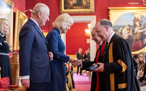 A man and woman in formal attire standing opposite two men, who are both dressed in academic robes. The woman is shaking hands with one of the men. They are stood inside a formal room painted dark red with large classic paintings on the walls.