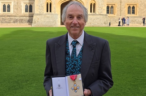 A man in a suit holding a box containing a medal, standing outside on green grass in front of a castle