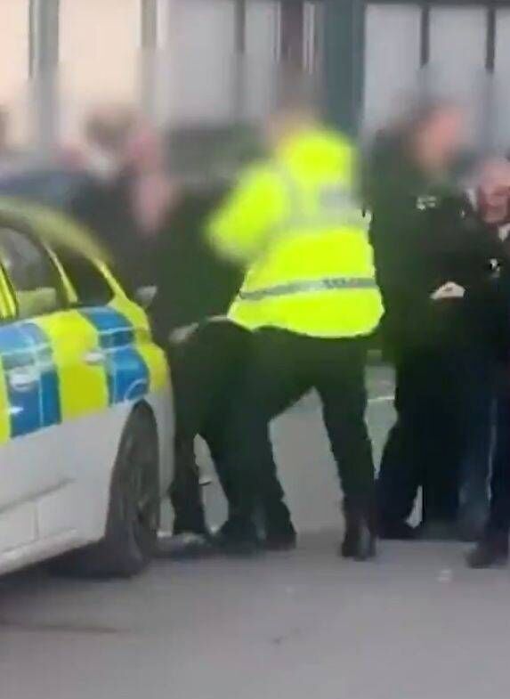 An image depicting a law enforcement officer in a reflective safety vest standing beside a police vehicle, with a group of indiv