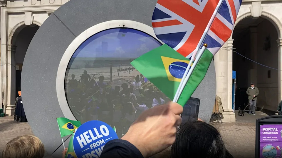 A circular "portal" showing an image of a crowd of people on a beach. People in front of the portal hold Brazilian and Union Jack flags