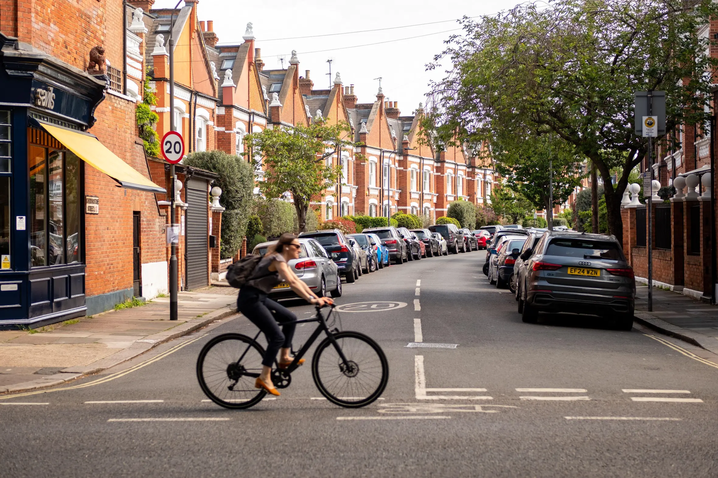 A woman cycling on New Kings Road in Parsons Green, London, lined with parked cars and shops.