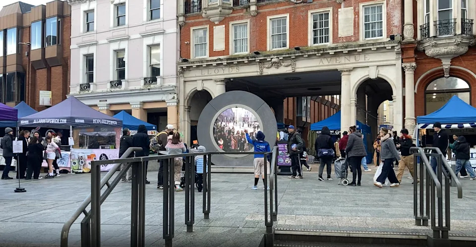 An open paved square in a town centre. A "portal" installation shows people in another country. People on the square are looking at it with one waving. An archway with "Lloyd's Avenue" written above it is behind the portal. There are a few gazebo stalls on the square, and railings in the foreground