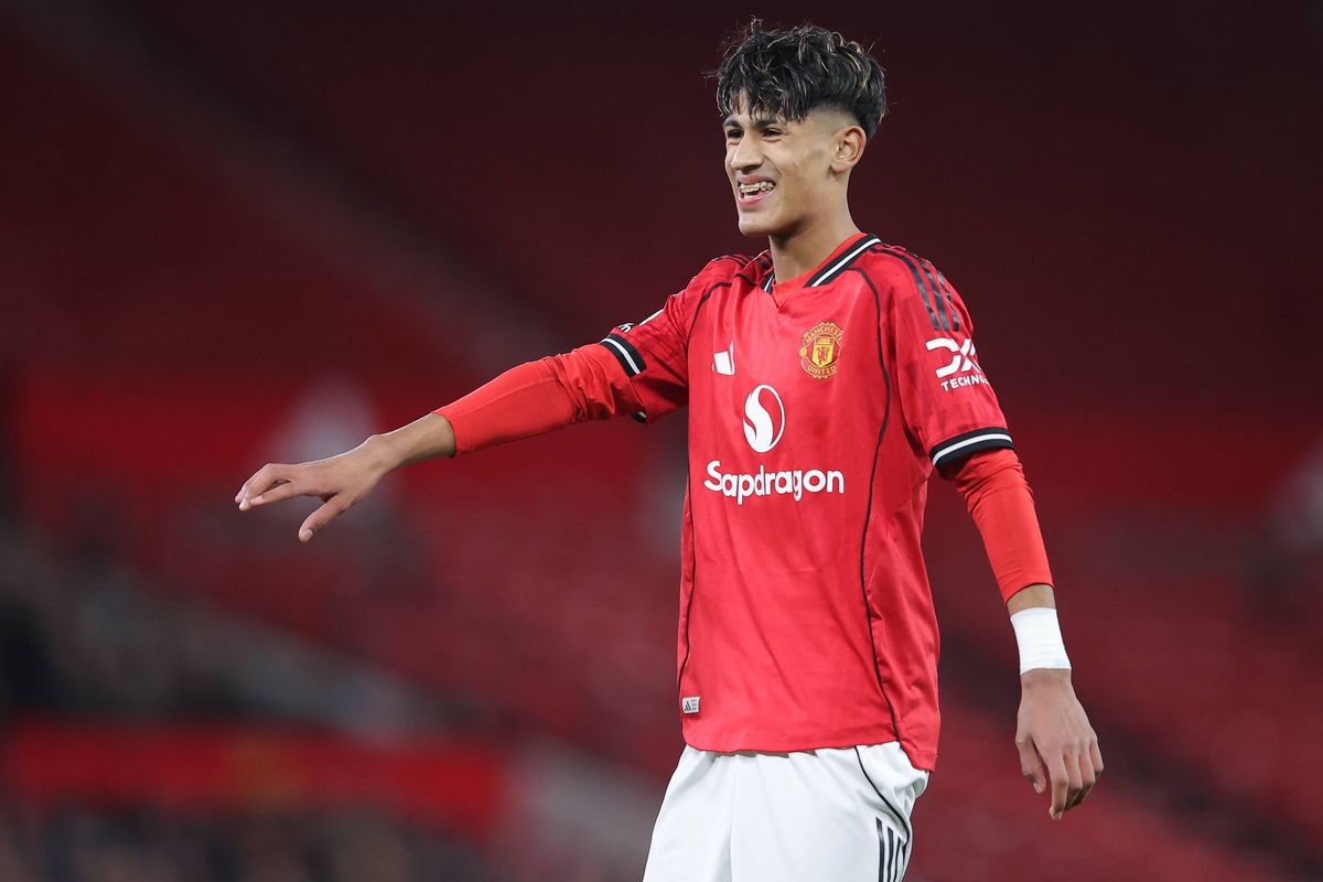 A male soccer player, wearing a red jersey with white accents and black trim, is captured mid-action on the field. He is stretching his right arm while his left arm is bent at the elbow. The player is positioned against a blurred red background, which could suggest an intense moment during a match. The jersey displays sponsor logos, including a prominent "S" logo and "Superson" text.
