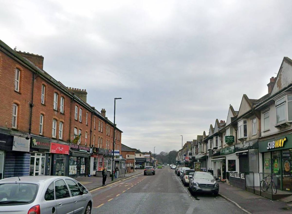 An urban street with several multi-story buildings on either side. The road features parked cars on the right side, and streetlights line the sidewalk. The sky appears overcast.