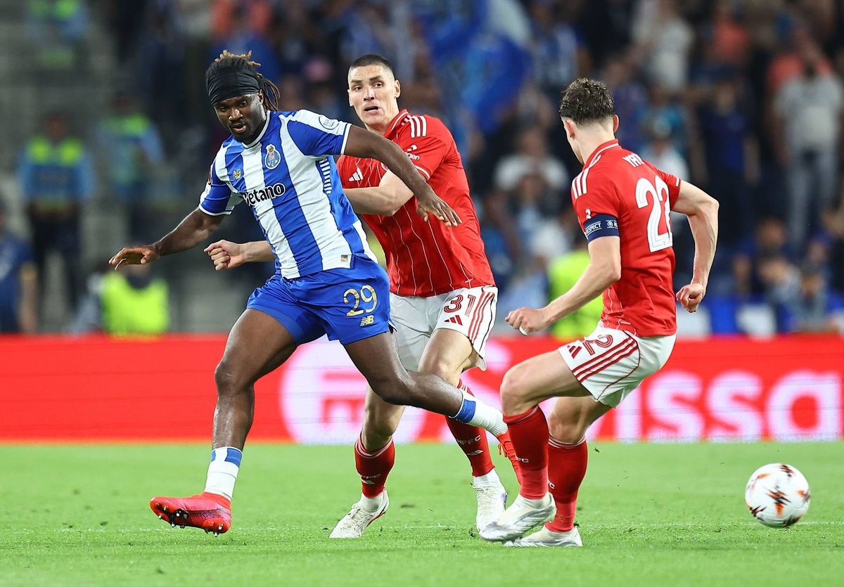 Three soccer players in uniform engage in a competitive match on the field, with one player in the foreground dribbling the ball while the other two attempt to intercept. The background features an audience seated in the stands, and the field is marked with sponsor logos.