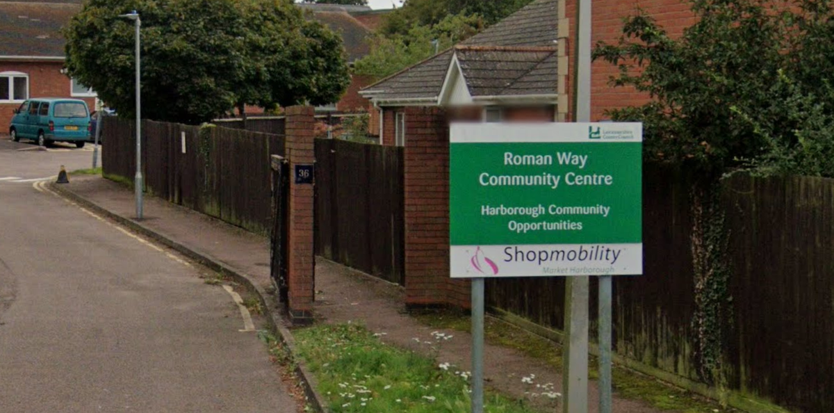 Sign depicting "Roman Way Community Centre, Harborough Community Opportunities" with white writing on a green background.  It has the Leicestershire County Council logo on it and at the bottom is the Shopmobility logo at the bottom with a white background. the sign is at the entrance to the community centre and is to the right of the picture.