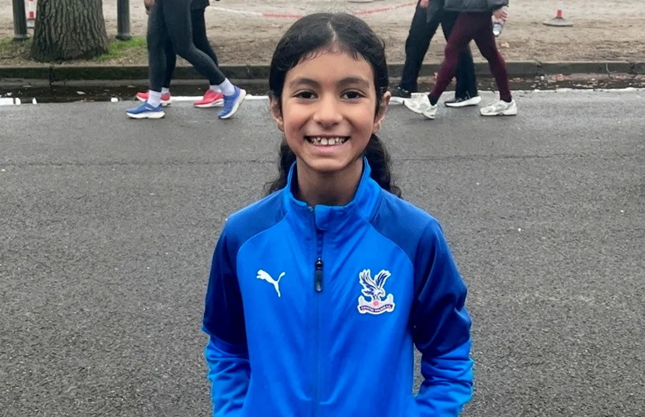 Nine-year-old Lily Janmohamed smiles at the camera, wearing a blue jacket, shorts, and white running shoes.