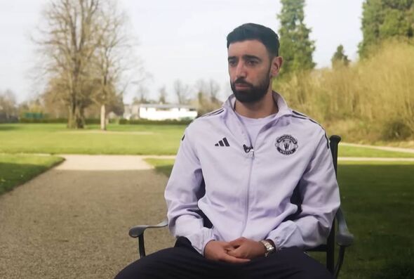 A man dressed in a white jacket and black pants sits on a chair in an outdoor setting, surrounded by a lush green lawn and trees