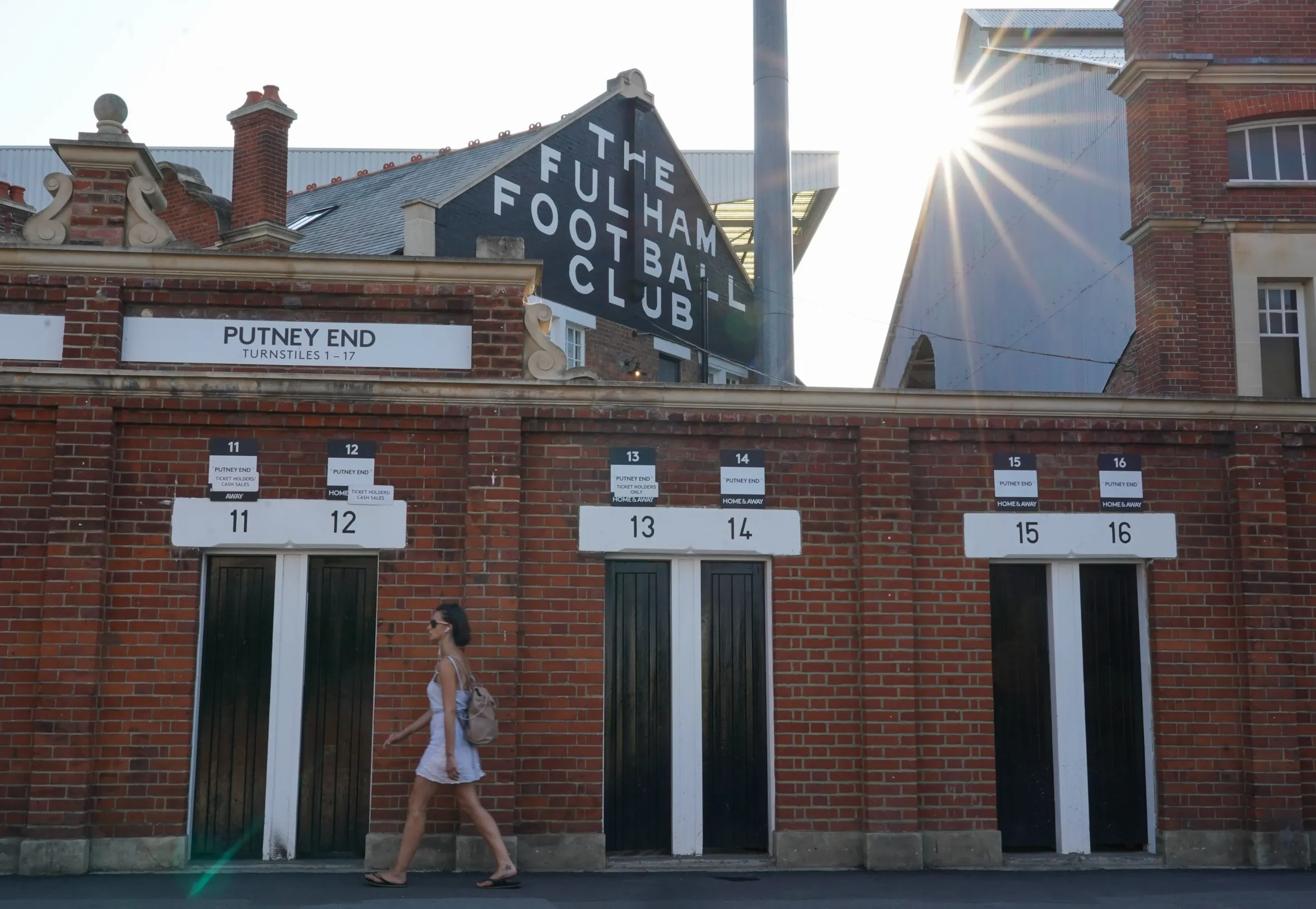 A woman walking past the brick wall of Craven Cottage, home of Fulham Football Club, with the low sun in the background.