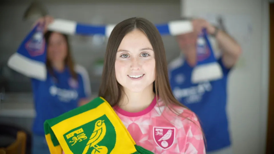 Eloise King has brown hair, and stands in front of her mother and father. She is wearing a pink Norwich City kit, and has a yellow and Green Norwich City scarf over her shoulder. 

Tracey King (left) and Chris King stand in the background wearing blue Ipswich Town shirts. They are holding a blue and white scarf between them.
