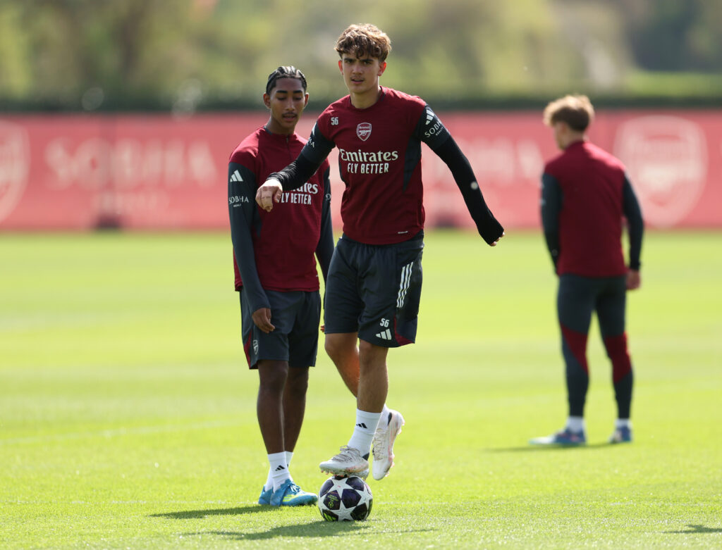 Max Dowman stands on the ball during Arsenal training. Marli Salmon stands behind him. They are both wearing Arsenal training kits of a deep red and blue shorts