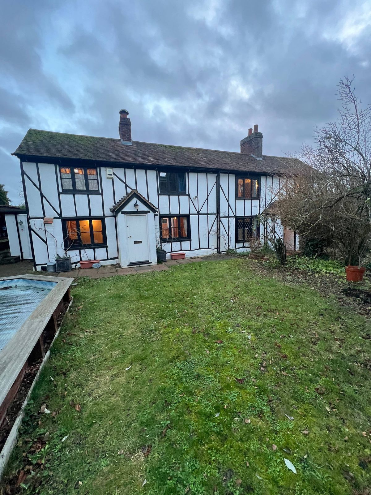 A traditional residential building with a thatched roof and white and black exterior walls. The property features a well-manicured lawn, a driveway, and several windows. The scene is set against a cloudy sky.