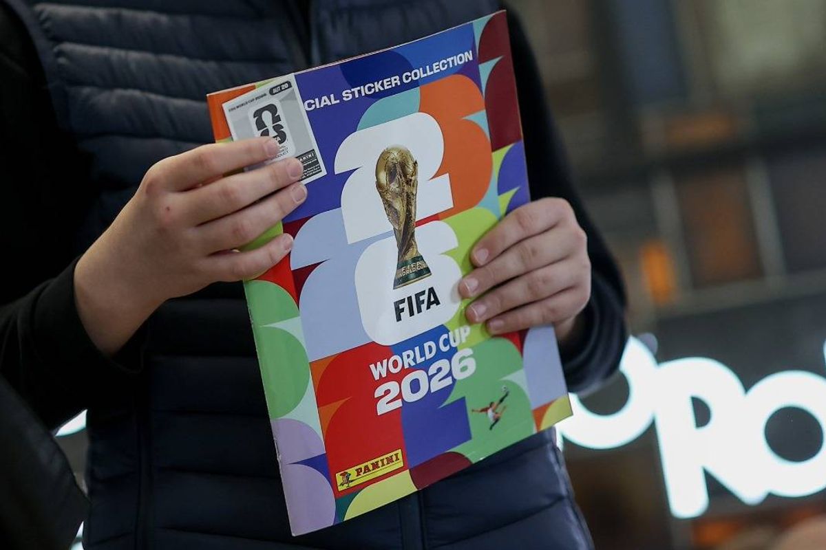 A person dressed in a dark jacket holds a colorful booklet featuring the FIFA World Cup 2026 logo, positioned against a blurred urban backdrop.