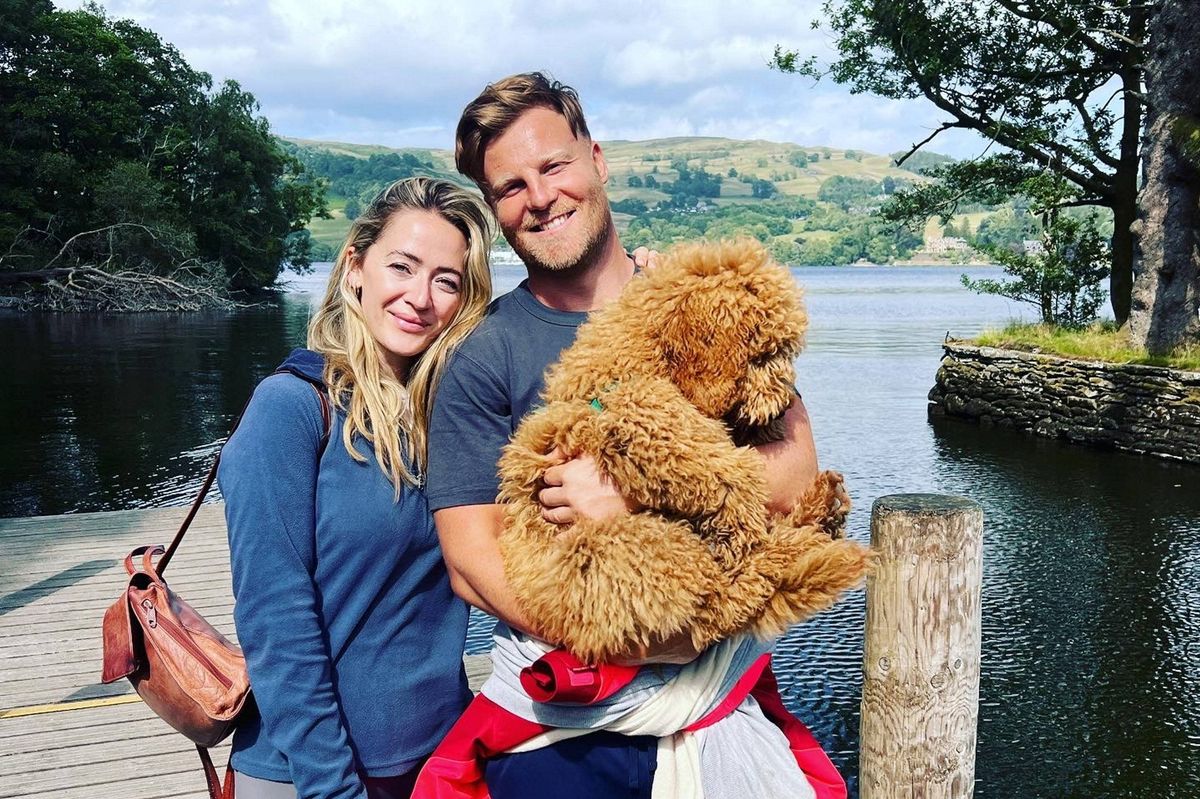 Two individuals are standing on a wooden pier by a lake, with one person holding a large brown teddy bear. They are both smiling and posing for a photograph. A tree and a body of water are visible in the background.